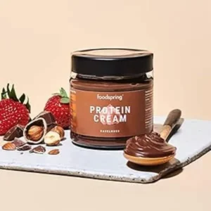 Crema proteica foodspring de avellanas con fresas, trozos de chocolate y cuchara sobre bandeja blanca