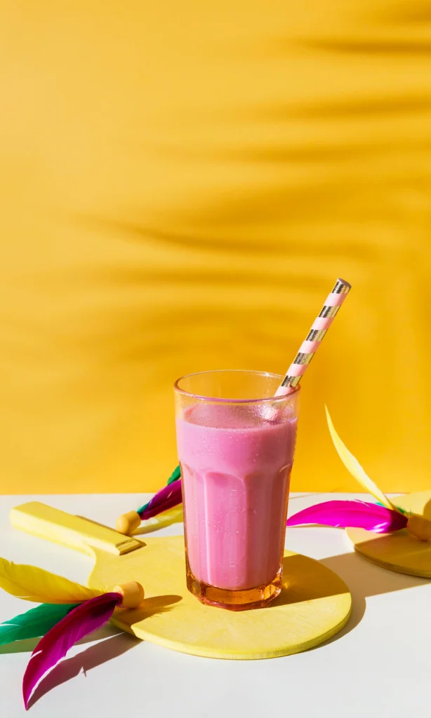 Vaso de batido rosa con pajita decorativa sobre fondo amarillo y elementos de colores vivos.