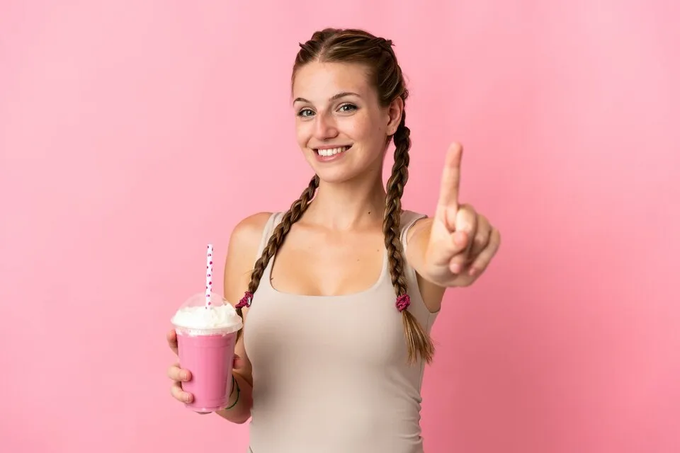 Mujer joven con dos trenzas sonríe mientras sostiene un batido rosa con nata y señala con el dedo índice hacia arriba, sobre fondo rosa.