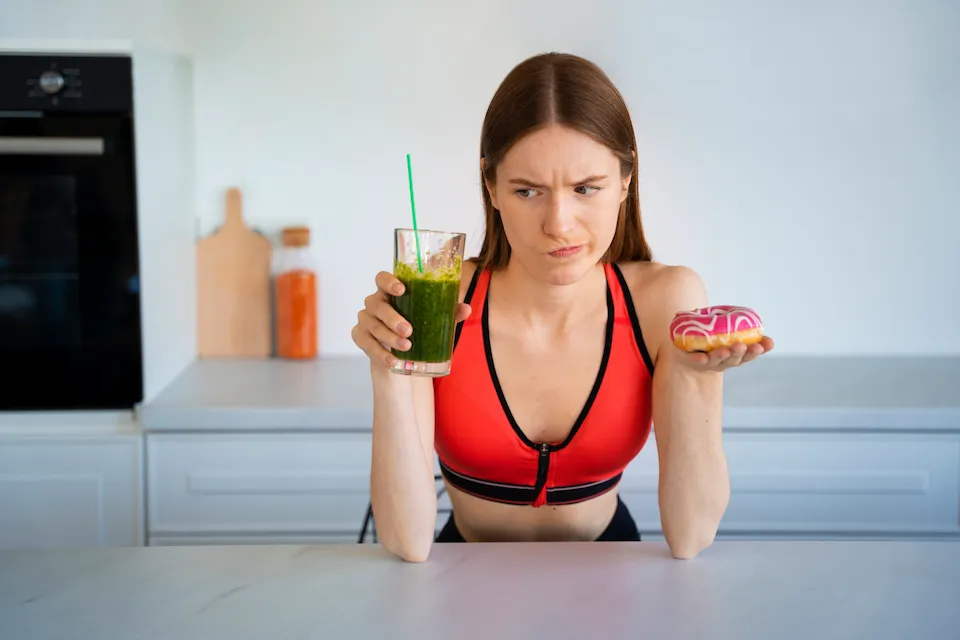 Mujer en ropa deportiva dudando entre un batido verde saludable y una dona con glaseado rosa.