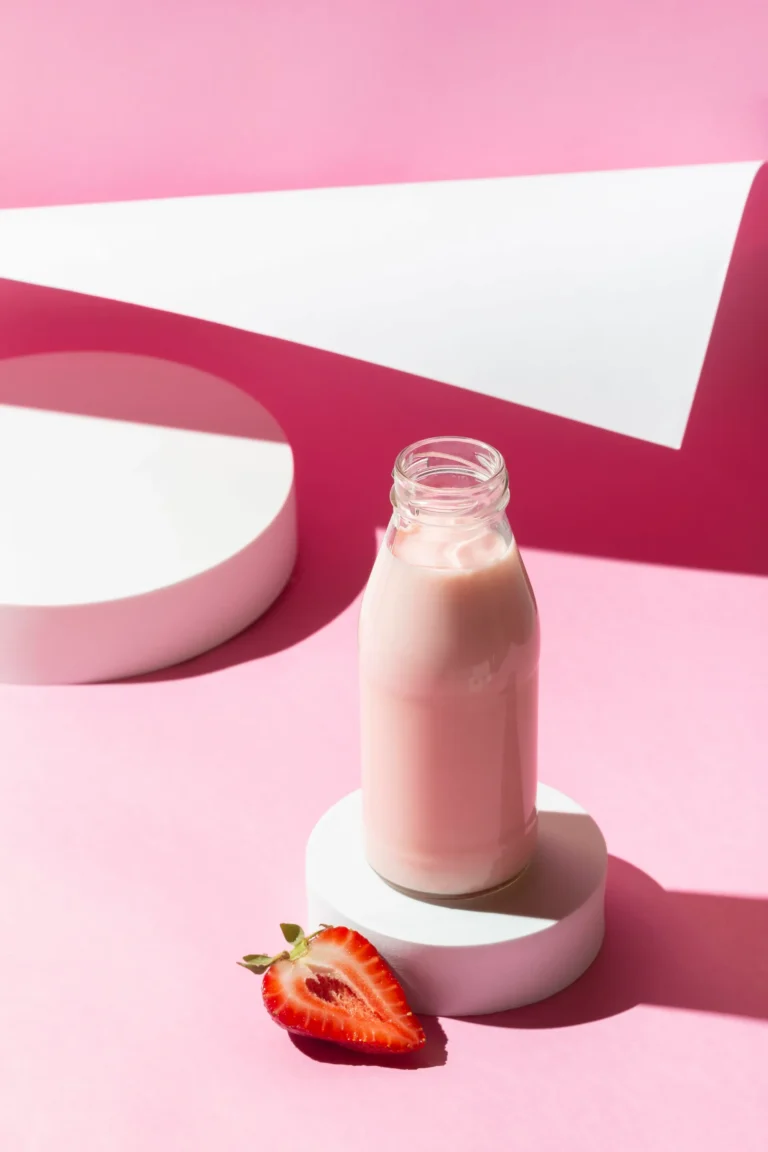 Batido sustitutivo de fresa listo para tomar en botella de cristal sobre fondo rosa, con media fresa fresca al lado.