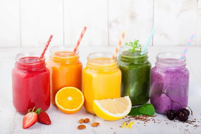 Cinco batidos de colores en tarros de cristal con pajita, acompañados de frutas frescas como fresas, naranja, limón, cerezas y hojas verdes, sobre fondo claro.