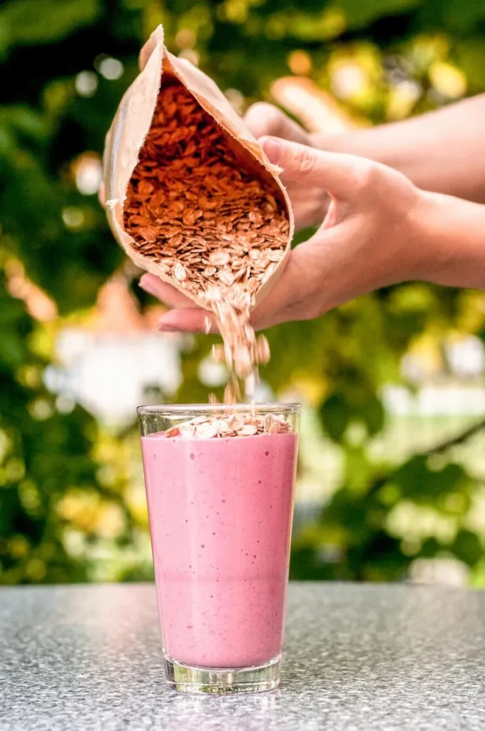 Persona vertiendo copos de avena sobre un batido sustitutivo rosa en un vaso de cristal, al aire libre